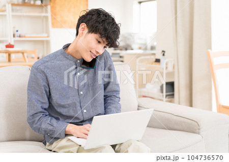 ソファーに座って膝の上にパソコンを置いてスマートフォンで電話している若い男性 ソファーに座って膝の上にパソコンを置いてスマートフォンで電話している若い男性 104736570