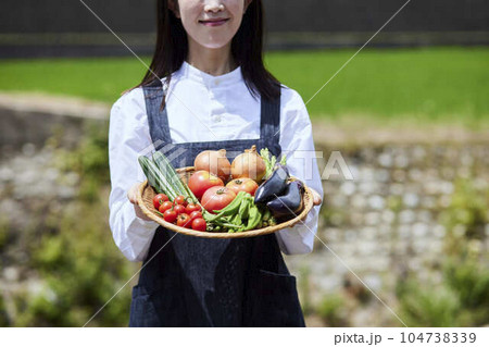 畑の前で採れたての野菜をもつ農家の女性 104738339