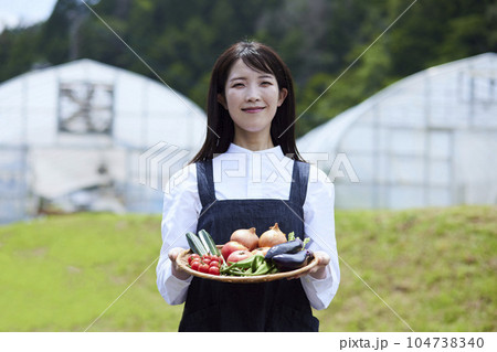 ビニールハウスの前で採れたての野菜をもつ農家の女性 104738340