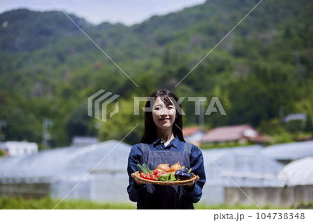 ビニールハウスの前で採れたての野菜をもつ農家の女性 104738348
