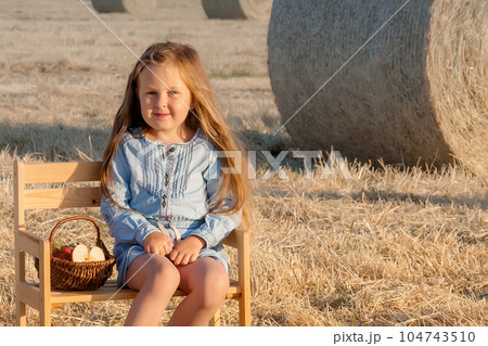 Cute girl sitting on a chair in the field, next to her is a basket of fresh fruit. Family summer fun in the country. Summer holidays. 104743510