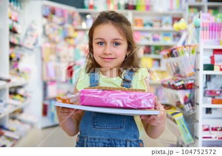 Caucasian little preschool girl collecting school supply and notebooks from stationery store, smiling looking at camera Caucasian little preschool girl collecting school supply and notebooks from stationery store, smiling looking at camera 104752572