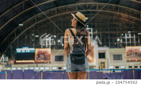 Rear view of young female traveller standing with backpack and hat at train station looking around for sign board to travel towards destination during vacation 104754502