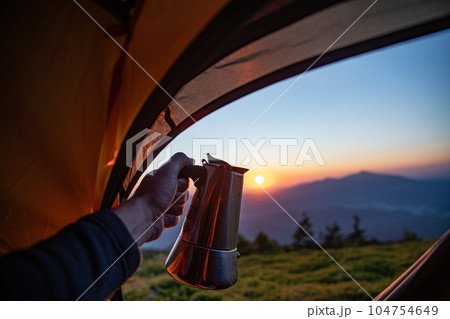 Man, a tourist, holds a device for making coffee - a moka pot on a gas burner in front of a tent on a mountain peak Man, a tourist, holds a device for making coffee - a moka pot on a gas burner in front of a tent on a mountain peak 104754649