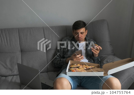 Man bachelor sits on sofa eating tasty pizza, drinking tea, browsing smartphone. 104755764