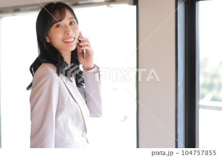 樹木の緑が見える窓辺で電話するロングヘアのビジネスウーマン_2 樹木の緑が見える窓辺で電話するロングヘアのビジネスウーマン_2 104757855
