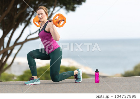 An adult woman trains with dumbbell on her shoulders - sports training outdoors near the sea 104757999