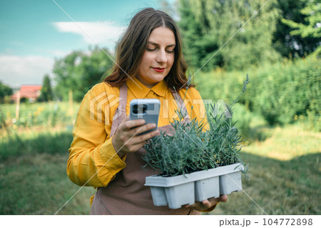 Phone call. Portrait of a young woman carrying flower pot with lavender while taking care of plants in the beautiful garden field. Caucasian farmer using a smartphone Phone call. Portrait of a young woman carrying flower pot with lavender while taking care of plants in the beautiful garden field. Caucasian farmer using a smartphone 104772898