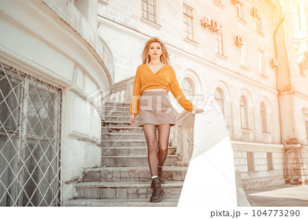 A middle-aged woman looks like a good blonde with curly beautiful hair and makeup on the background of the building. She is wearing a yellow sweater. 104773290