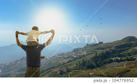 Father and son over meadowputs his son on his shoulder. The family life journey concept Father and son over meadowputs his son on his shoulder. The family life journey concept 104776952