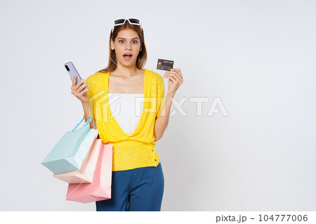Excited young woman carrying shopping bags with using credit card and mobile phone standing isolated on white background. 104777006
