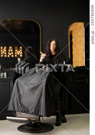 Beautiful young brutal woman male master hairdresser, barber holding razor for shaving standing near chair in barbershop Beautiful young brutal woman male master hairdresser, barber holding razor for shaving standing near chair in barbershop 104780996