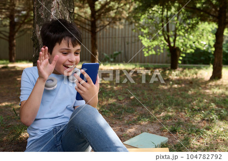 Cheerful teen boy waving hello with hand, having online conference by video link on his smartphone, sitting under a tree Cheerful teen boy waving hello with hand, having online conference by video link on his smartphone, sitting under a tree 104782792