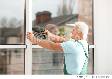 Mature construction worker repairing plastic window with electric screwdriver indoors 104786639