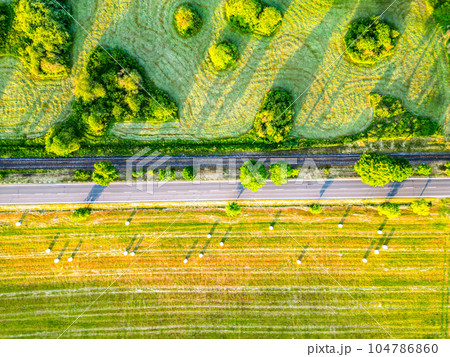 Asphalt road and concurrent railway in lush green morning lagricultural landscape. Aerial view from drone. Asphalt road and concurrent railway in lush green morning lagricultural landscape. Aerial view from drone. 104786860
