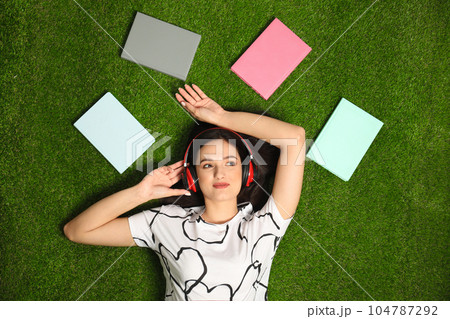 Young woman listening to audiobook while lying on grass, flat lay 104787292