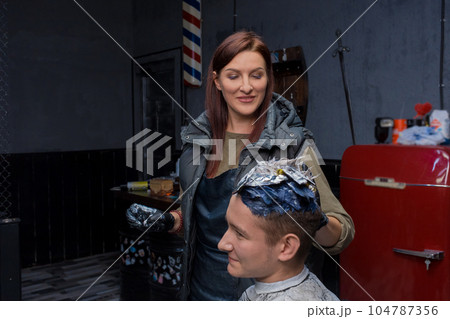 Smiling positive brunette, girl, professional barbershop worker at work, dyeing hair to a client, a young guy, parking services Smiling positive brunette, girl, professional barbershop worker at work, dyeing hair to a client, a young guy, parking services 104787356