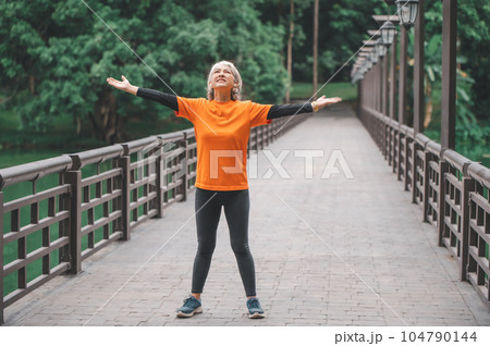 elderly white-haired Asian woman spreading her hands to breathe in the fresh air while exercising in the park early in the morning. 104790144