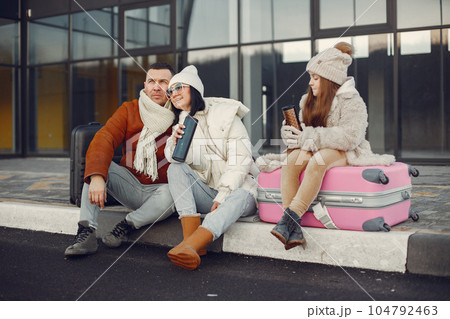 Family of three going on vacation. Man and girl sitting on a luggage and drinking tea from thermos. Mother, father and daughter wearing warm clothes. Family of three going on vacation. Man and girl sitting on a luggage and drinking tea from thermos. Mother, father and daughter wearing warm clothes. 104792463