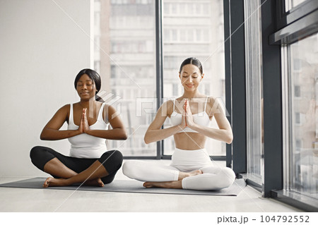 Two women stretching on yoga mat in bright studio with big windows. Caucasian woman wearing white leggins and top and black woman white t-shirt and black leggins. Sport, fitness, diet, nutrition and Two women stretching on yoga mat in bright studio with big windows. Caucasian woman wearing white leggins and top and black woman white t-shirt and black leggins. Sport, fitness, diet, nutrition and 104792552