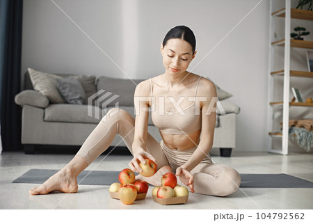 Slim and beautiful woman wearing beige leggins and top and sitting on yoga mat. Young girl with fit body posing at home with fresh apples. Sport, fitness, diet, nutrition and health care concept. Slim and beautiful woman wearing beige leggins and top and sitting on yoga mat. Young girl with fit body posing at home with fresh apples. Sport, fitness, diet, nutrition and health care concept. 104792562
