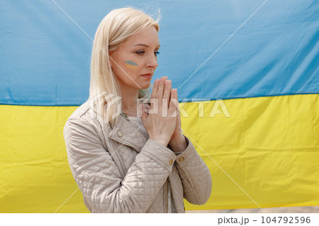 Woman with ukrainian flag on a cheek standing in front of big flag of Ukraine. Girl praying. Girl supporting Ukraine. Woman with ukrainian flag on a cheek standing in front of big flag of Ukraine. Girl praying. Girl supporting Ukraine. 104792596