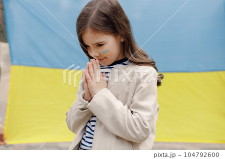 Little girl with ukrainian flag on a cheek standing in front of big flag of Ukraine. Young pretty girl praying. Girl supporting Ukraine. 104792600
