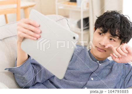 リビングのソファーで寝転がってタブレット端末で動画を見て泣く若い男性 リビングのソファーで寝転がってタブレット端末で動画を見て泣く若い男性 104793885