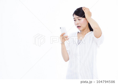 白い背景の前でスマホを持ち頭を抱えて驚く30代の女性 白い背景の前でスマホを持ち頭を抱えて驚く30代の女性 104794586