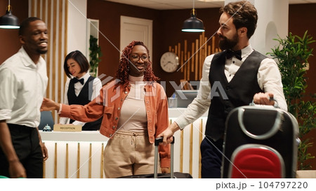Happy african american tourists tipping helpful bellboy carrying their luggage in resort lobby. Cheerful guests checking out from hotel after handing over room access key card to friendly receptionist 104797220