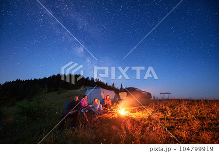 Family camp in mountains in the evening. Group of tourists having a rest near campfire on a top of hill, near tourist tent and off-road vehicle. Starry night in Carpathian mountains. 104799919
