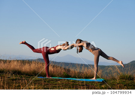 Two girls doing yoga pose at early morning. Sporty mother and her daughter exercising outside. Outdoor meditation in mountains. Two girls doing yoga pose at early morning. Sporty mother and her daughter exercising outside. Outdoor meditation in mountains. 104799934