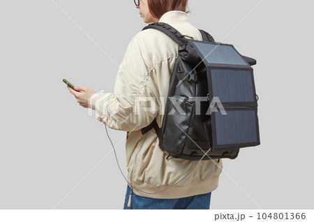 Young woman carrying backpack with flexible solar panel attached to smartphone in her hands 104801366
