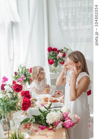 A little blonde girl with her mom on a kitchen countertop decorated with peonies. The concept of the relationship between mother and daughter. Spring atmosphere. A little blonde girl with her mom on a kitchen countertop decorated with peonies. The concept of the relationship between mother and daughter. Spring atmosphere. 104802054