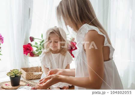 A little blonde girl with her mom on a kitchen countertop decorated with peonies. The concept of the relationship between mother and daughter. Spring atmosphere. A little blonde girl with her mom on a kitchen countertop decorated with peonies. The concept of the relationship between mother and daughter. Spring atmosphere. 104802055