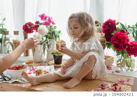 A little blonde girl on a kitchen countertop decorated with peonies. Spring atmosphere. A little blonde girl on a kitchen countertop decorated with peonies. Spring atmosphere. 104802421