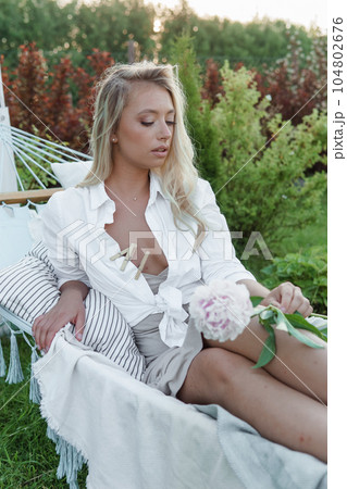 A beautiful blonde woman is lying on a hammock in her summer cottage. The concept of life in the country and summer holidays. A beautiful blonde woman is lying on a hammock in her summer cottage. The concept of life in the country and summer holidays. 104802676