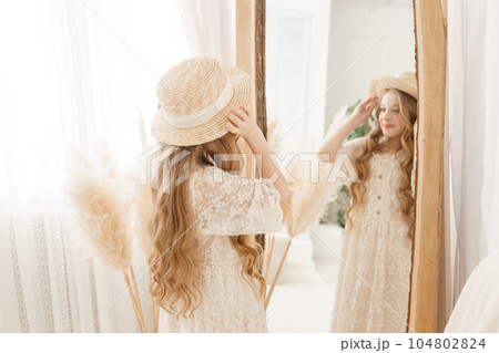A beautiful teenage girl with long hair measures a straw hat in front of a mirror. Self-admiration of a blonde. 104802824