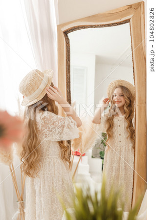 A beautiful teenage girl with long hair measures a straw hat in front of a mirror. Self-admiration of a blonde. 104802826