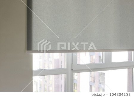 Details of gray fabric roller blinds. Automatic curtain close-up on the window. Selective focus. Motorized shades. City outside the window. Details of gray fabric roller blinds. Automatic curtain close-up on the window. Selective focus. Motorized shades. City outside the window. 104804152