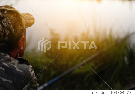A man is engaged in fishing by a lake or pond, using a rod and spinning reel. He stands on the river bank surrounded by natural scenery. The image offers copy space for various design purposes 104804378