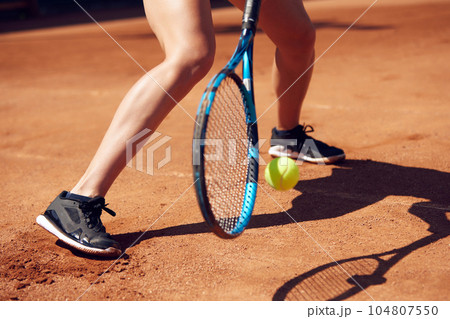 Cropped image of female legs in sportswear and tennis racket with ball over floor background with shadows. Open air tennis court Cropped image of female legs in sportswear and tennis racket with ball over floor background with shadows. Open air tennis court 104807550