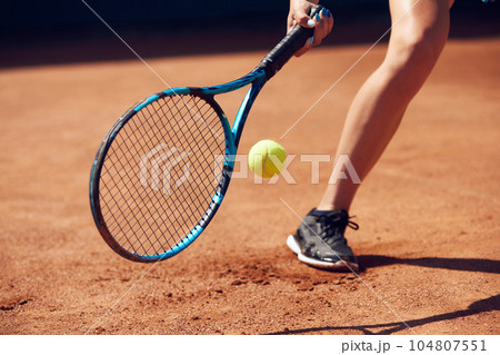 Cropped image of female legs in sportswear and tennis racket with ball over floor background with shadows. Open air tennis court 104807551