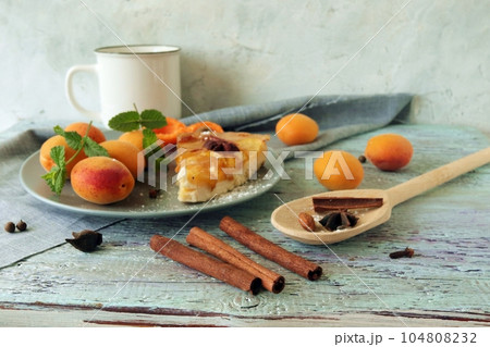 A piece of apricot pie on a plate, sprinkled with powdered sugar, spices and mint leaves, fresh apricots on the kitchen table, homemade food, sweet dessert 104808232