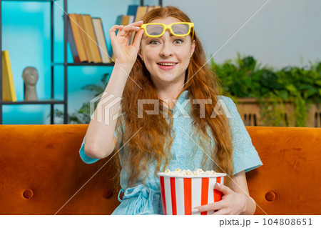 Woman sitting on couch eating popcorn and watching interesting TV 3D film, sport game online at home 104808651