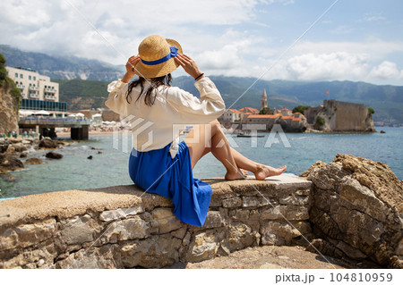 Montenegro, Budva. A girl in a blue dress sits on stones and looks at the central square in Budva, Montenegro.Selective focus, noise. Vacation concept, travel to Europe. 104810959