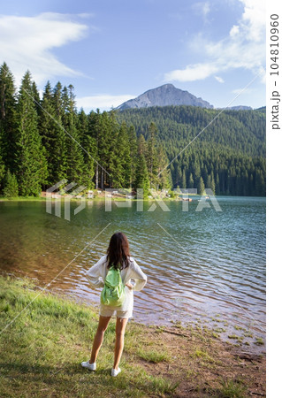 A girl with a bright backpack stands with her back near a mountain lake in the background. Positive young woman traveling on a blue lake outdoors. The girl admires the view. 104810960