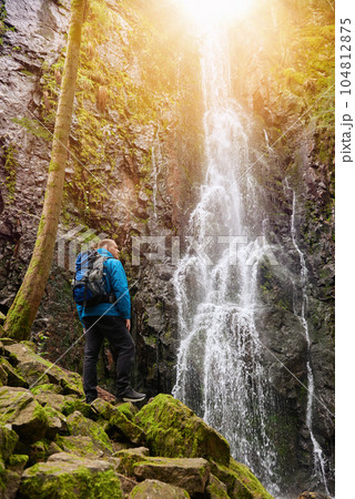 Tourist attraction of Germany - falls of Burgbach Waterfall near Schapbach, Black Forest, Baden-Wurttemberg, Germany. Man hiker in blue jacket standing on stone and looks at flow of falling water 104812875