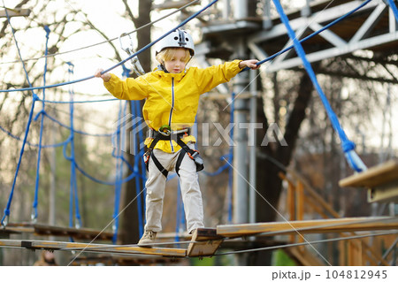 Little boy having fun in Adventure Park for children amoung ropes, stairs, bridges. Outdoor climbing adventure playground in public park. Activity entertainment for kids Little boy having fun in Adventure Park for children amoung ropes, stairs, bridges. Outdoor climbing adventure playground in public park. Activity entertainment for kids 104812945