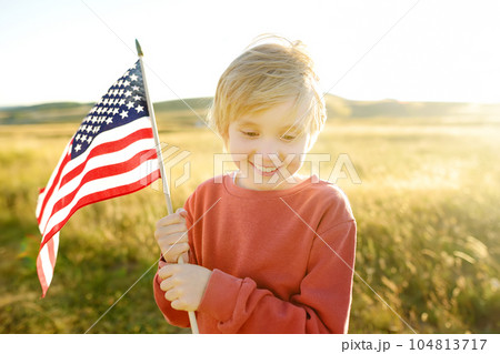 Cute little boy celebrating of July, 4 Independence Day of USA at sunny summer sunset. Child running with american flag of United States on wheat field. Proud small american boy holding country flag Cute little boy celebrating of July, 4 Independence Day of USA at sunny summer sunset. Child running with american flag of United States on wheat field. Proud small american boy holding country flag 104813717